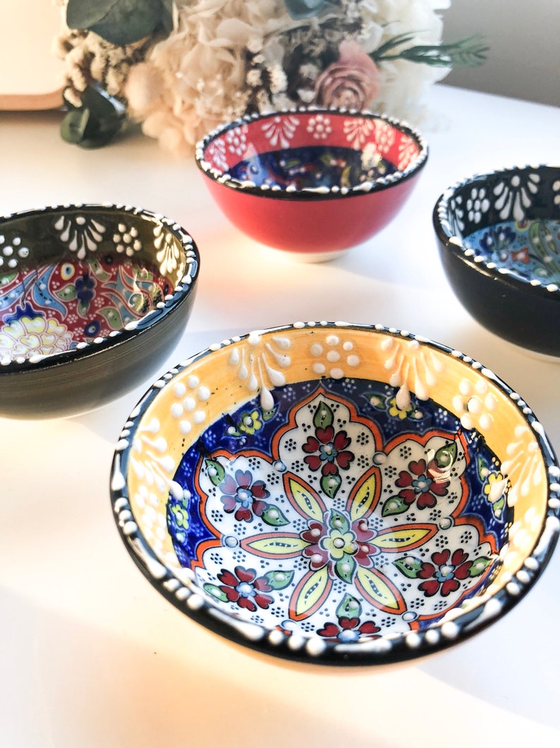 May include: Four small, colorful ceramic bowls with intricate floral patterns. The bowls have a variety of colors, including red, black, yellow, and blue. The interior designs feature detailed floral motifs, and the rims are decorated with white dots.