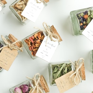 May include: A collection of small glass jars filled with various dried tea blends. Each jar is tied with twine and a small tag that reads "Let Love Brew" with the couple's names, "Bryan & Katie", and the type of tea.