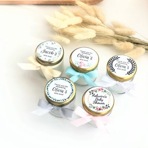 May include: Five small glass jars with gold lids and colorful labels. The labels have floral designs and text that says "Thank you for showering [name]'s with love!" and "Ashwini's Baby Shower".