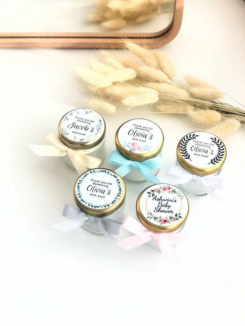 May include: Five small glass jars with gold lids and colorful labels. The labels have floral designs and text that says "Thank you for showering [name]'s with love!" and "Ashwini's Baby Shower".