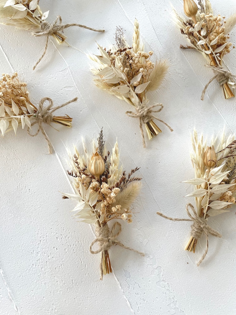 May include: Six small bouquets of dried flowers tied with twine. The bouquets are made of dried grasses, flowers, and seed pods in shades of beige and brown. The bouquets are arranged on a white surface.