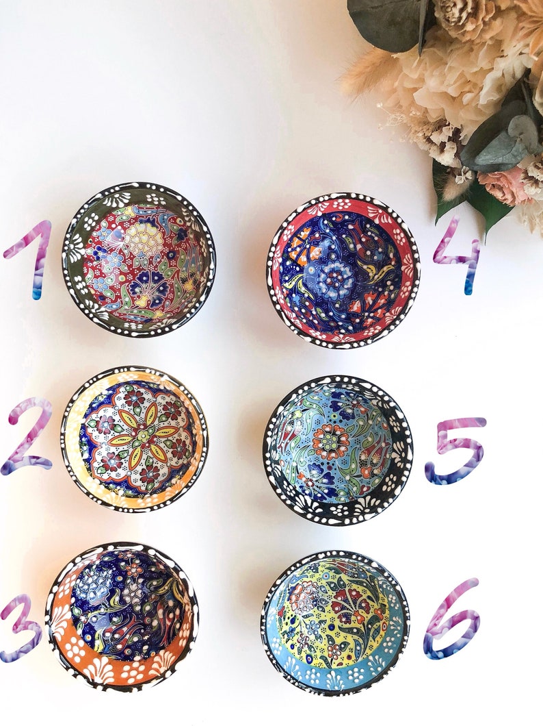 May include: Six small, round ceramic bowls with intricate floral patterns in various colors. The bowls have black rims with white dots and are arranged on a white surface. The bowls feature red, blue, green, yellow, and orange designs.