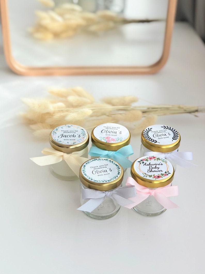 May include: Five small glass jars with gold lids and floral themed labels. The labels have text that says "Thank you for showering" followed by a name, and "with love". One jar has a label that says "Ashwini's Baby Shower".