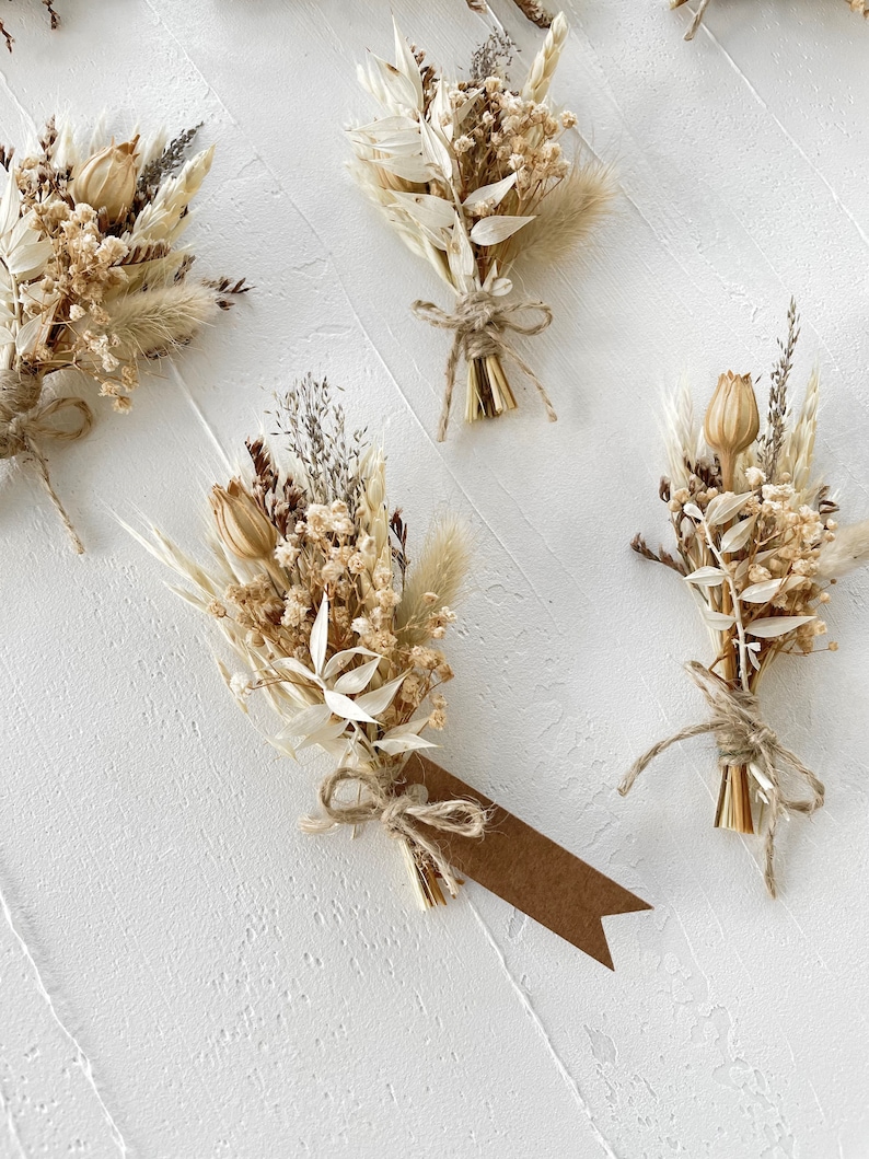 May include: Four small bouquets of dried flowers tied with twine. The bouquets are made of dried grasses, white flowers, and small brown seed pods. Each bouquet has a small brown paper tag attached.