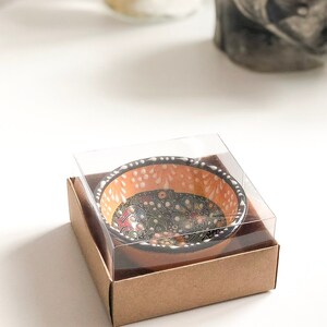 May include: A small, decorative ceramic bowl with a floral pattern in a clear plastic case and a brown cardboard box. The bowl has an orange interior with white dots and a black rim. The floral design includes red, yellow, and blue accents.