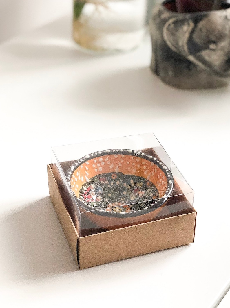 May include: A small, decorative ceramic bowl with a floral pattern in a clear plastic case and a brown cardboard box. The bowl has an orange interior with white dots and a black rim. The floral design includes red, yellow, and blue accents.