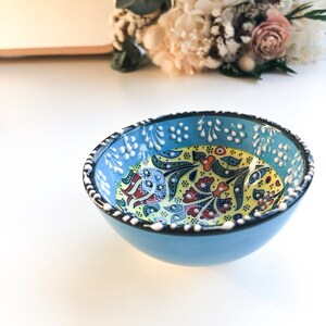 May include: A small, round, light blue ceramic bowl with a detailed floral pattern in yellow, red, and blue. The rim is decorated with white dots. The bowl is likely handmade and suitable for serving small portions.