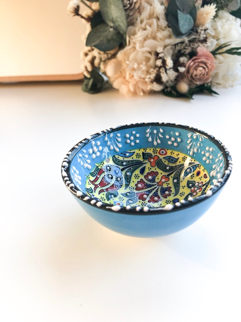 May include: A small, round, light blue ceramic bowl with a detailed floral pattern in yellow, red, and blue. The rim is decorated with white dots. The bowl is likely handmade and suitable for serving small portions.