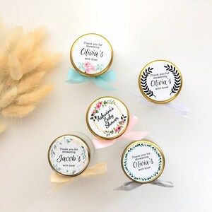 May include: Five small glass jars with gold lids and white labels. The labels have floral designs and text that says "Thank you for showering Olivia's with love", "Ashwini's Baby Shower", "Thank you for showering Jacob's with love", and "Thank you for showering Olivia's with love".