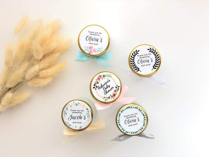 May include: Five small glass jars with gold lids and white labels. The labels have floral designs and text that says "Thank you for showering Olivia's with love", "Ashwini's Baby Shower", "Thank you for showering Jacob's with love", and "Thank you for showering Olivia's with love".