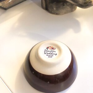 May include: A small, ceramic bowl with a two-tone design: a dark brown base and a cream-colored upper section. The bottom of the bowl features a sticker with the text "Stanton Wedding March 23, 2019" surrounded by floral illustrations. The bowl is on a white surface.