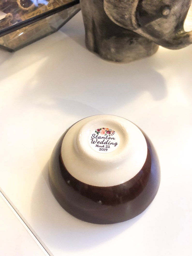 May include: A small, ceramic bowl with a two-tone design: a dark brown base and a cream-colored upper section. The bottom of the bowl features a sticker with the text "Stanton Wedding March 23, 2019" surrounded by floral illustrations. The bowl is on a white surface.