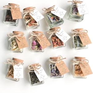 May include: A collection of small glass jars with cork lids, each filled with dried flowers, herbs, and tea. The jars are tied with twine and have small brown paper tags with the text "Let Love Bloom Bryan & Katie" printed on them.