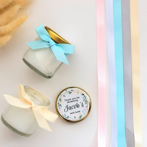 May include: Two small glass jars with white cream inside, tied with blue and cream ribbons. The jars are on a white surface with a gold lid and a sticker that says "Thank you for showering Jacob's with love".