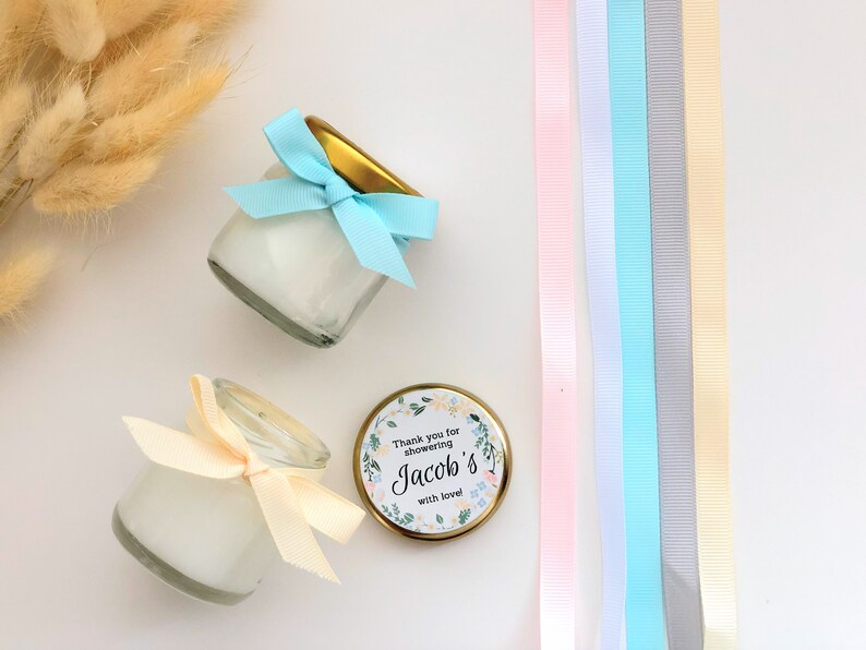 May include: Two small glass jars with white cream inside, tied with blue and cream ribbons. The jars are on a white surface with a gold lid and a sticker that says "Thank you for showering Jacob's with love".
