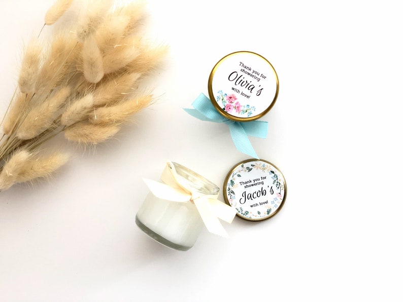 May include: Two small glass jars with white candles and gold lids. The lids have floral patterned labels with the text "Thank you for showering Olivia's with love!" and "Thank you for showering Jacob's with love!" The jars are tied with blue and white ribbons.