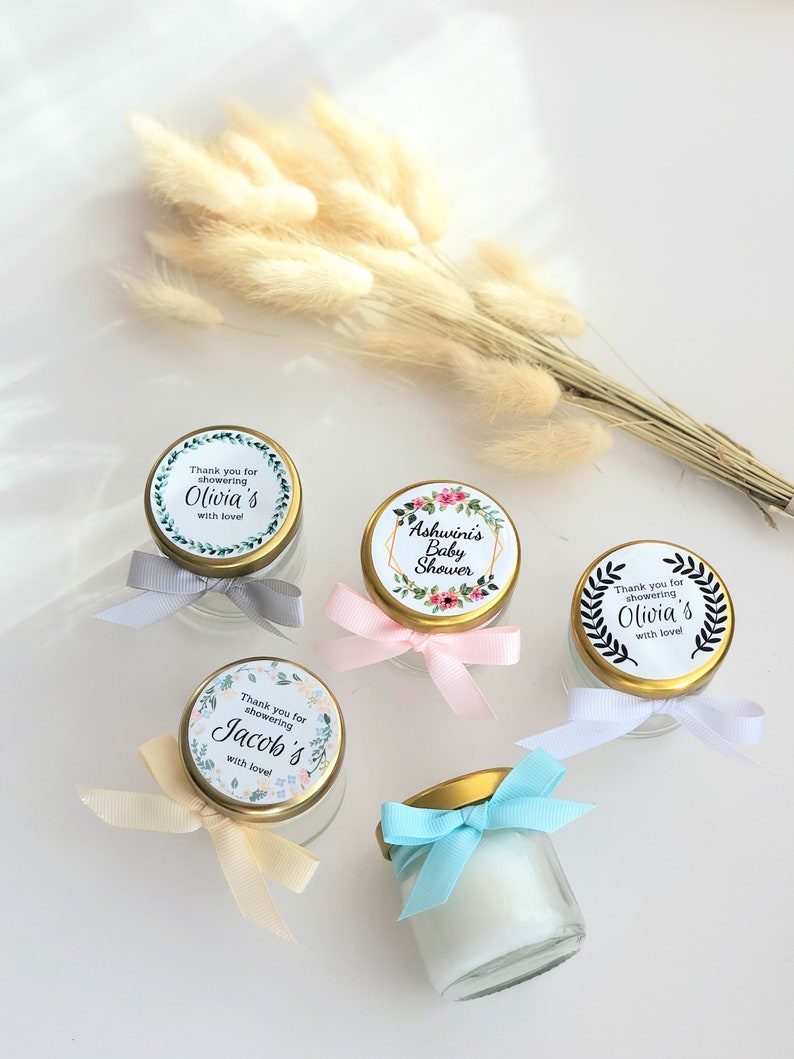 May include: Four small glass jars with gold lids and colorful ribbons. Each jar has a round label with a floral design and text that says "Thank you for showering [name]'s with love!" The names on the labels are Olivia, Olivia, Jacob, and Ashwini.