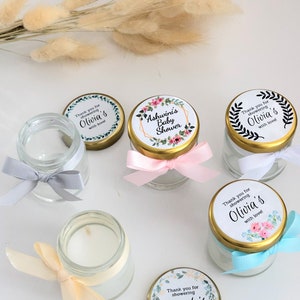 May include: Assortment of small glass jars with lids, each decorated with a floral design and personalized text. The jars are tied with ribbons in various colors, including pink, blue, and cream. The text on the lids reads "Thank you for showering Olivia's with love!" and "Ashwini's Baby Shower".