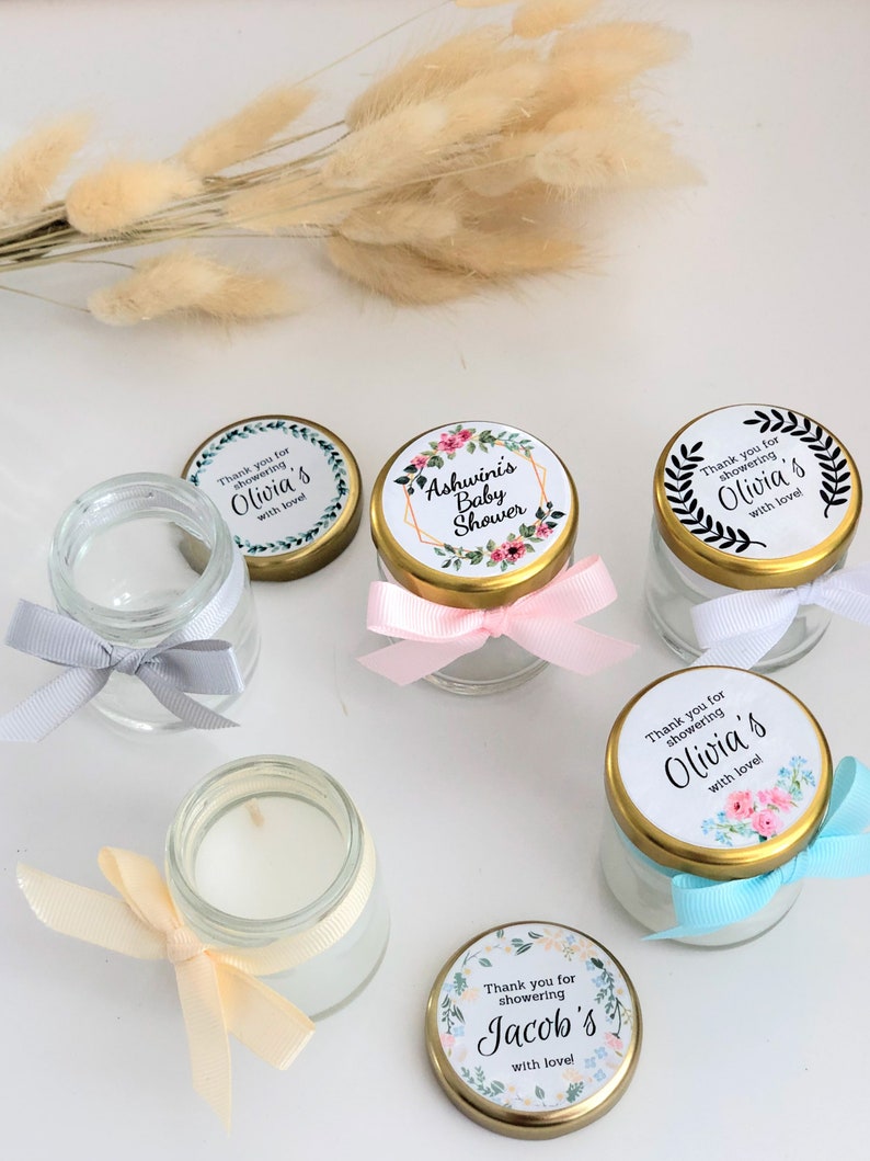 May include: Assortment of small glass jars with lids, each decorated with a floral design and personalized text. The jars are tied with ribbons in various colors, including pink, blue, and cream. The text on the lids reads "Thank you for showering Olivia's with love!" and "Ashwini's Baby Shower".