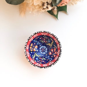 May include: A small, round ceramic bowl with a vibrant floral design. The bowl features a deep blue interior with orange and white floral patterns. The rim is red with white dots and a black border. Decorative and colorful.