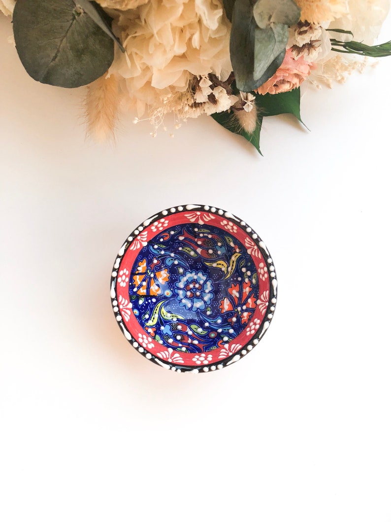 May include: A small, round ceramic bowl with a vibrant floral design. The bowl features a deep blue interior with orange and white floral patterns. The rim is red with white dots and a black border. Decorative and colorful.