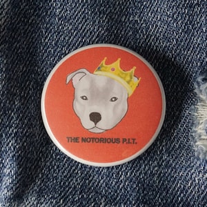 May include: A round, red button with a white border featuring a pit bull wearing a gold crown. The text "THE NOTORIOUS P.I.T." is printed below the dog. The button is on a denim jacket.