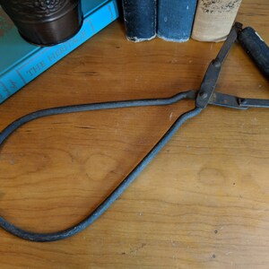 May include: A vintage metal log grabber with a wooden handle. The grabber is made of black metal and has a long, curved handle.