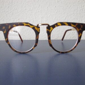 Puede incluir: Gafas con montura con estampado de carey. Las gafas tienen una forma única con la parte superior recta e inferior redondeada. Las lentes son transparentes y las patillas son de color marrón oscuro. Sobre una superficie azul.