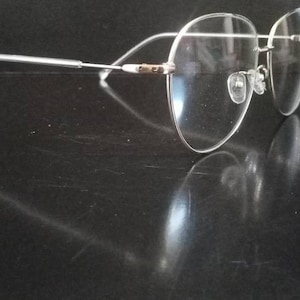Puede incluir: Un par de gafas transparentes sin montura con brazos de metal plateado. Las gafas están sobre una superficie negra.
