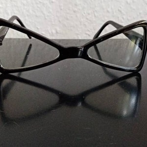 May include: Black cat-eye glasses with a thin frame and clear lenses. The glasses are lying on a black surface.
