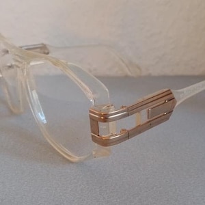Puede incluir: Gafas transparentes con montura rectangular y detalles plateados en las patillas. Las gafas están sobre una superficie azul claro, con un fondo blanco.