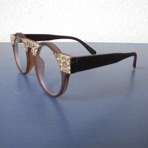 Puede incluir: Gafas de plástico marrón con detalles dorados en la barra del frente y las patillas. Las lentes son redondas.