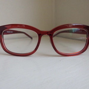 Könnte beinhalten: Rote Kunststoff-Brille mit klaren Gläsern. Die Brille hat eine Cat-Eye-Form.