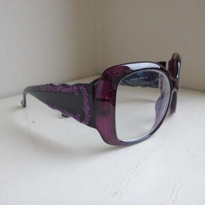 Puede incluir: Un par de gafas de gran tamaño con montura morada oscura y lentes transparentes. La montura tiene una base negra con un patrón decorativo morado en las patillas. Las gafas están sobre un fondo blanco.