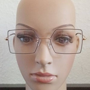 Könnte beinhalten: Eine Brille mit goldfarbenem Rahmen, klaren Gläsern und einem geometrischen, eckigen Gestell.