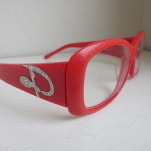 Puede incluir: Gafas de plástico rojo con lentes transparentes. Las monturas tienen un diseño plateado con la letra "P" repetida.