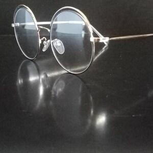 Puede incluir: Gafas redondas de montura plateada con lentes transparentes. Las gafas están sobre una superficie negra y se ve el reflejo de las gafas.