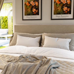 May include: A beige upholstered bed with a white duvet and a beige throw blanket. Two framed prints with the text "Fleurs &Eacute;panouies" hang above the headboard.