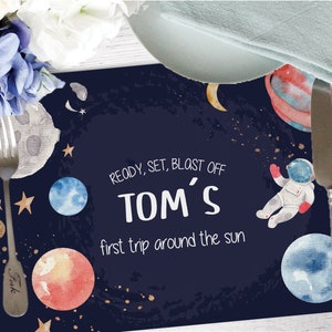 May include: A blue and black space-themed placemat featuring planets, stars, and an astronaut. The text reads "Ready, Set, Blast Off Tom's First Trip Around The Sun".