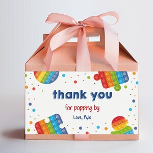 May include: A pink favor box with a pink ribbon and a white label that says "thank you for popping by Love, Kyle" with colorful pop it toys on the label.