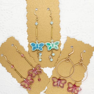 May include: Three pairs of butterfly earrings on gold display cards. The first pair is turquoise and blue with crystal accents. The second pair is pink with crystal accents. The third pair is pink with gold hoops.