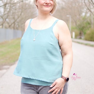 May include: A woman wearing a light blue sleeveless top with a silver necklace and a silver heart pendant. She is also wearing gray jeans and a brown watch.