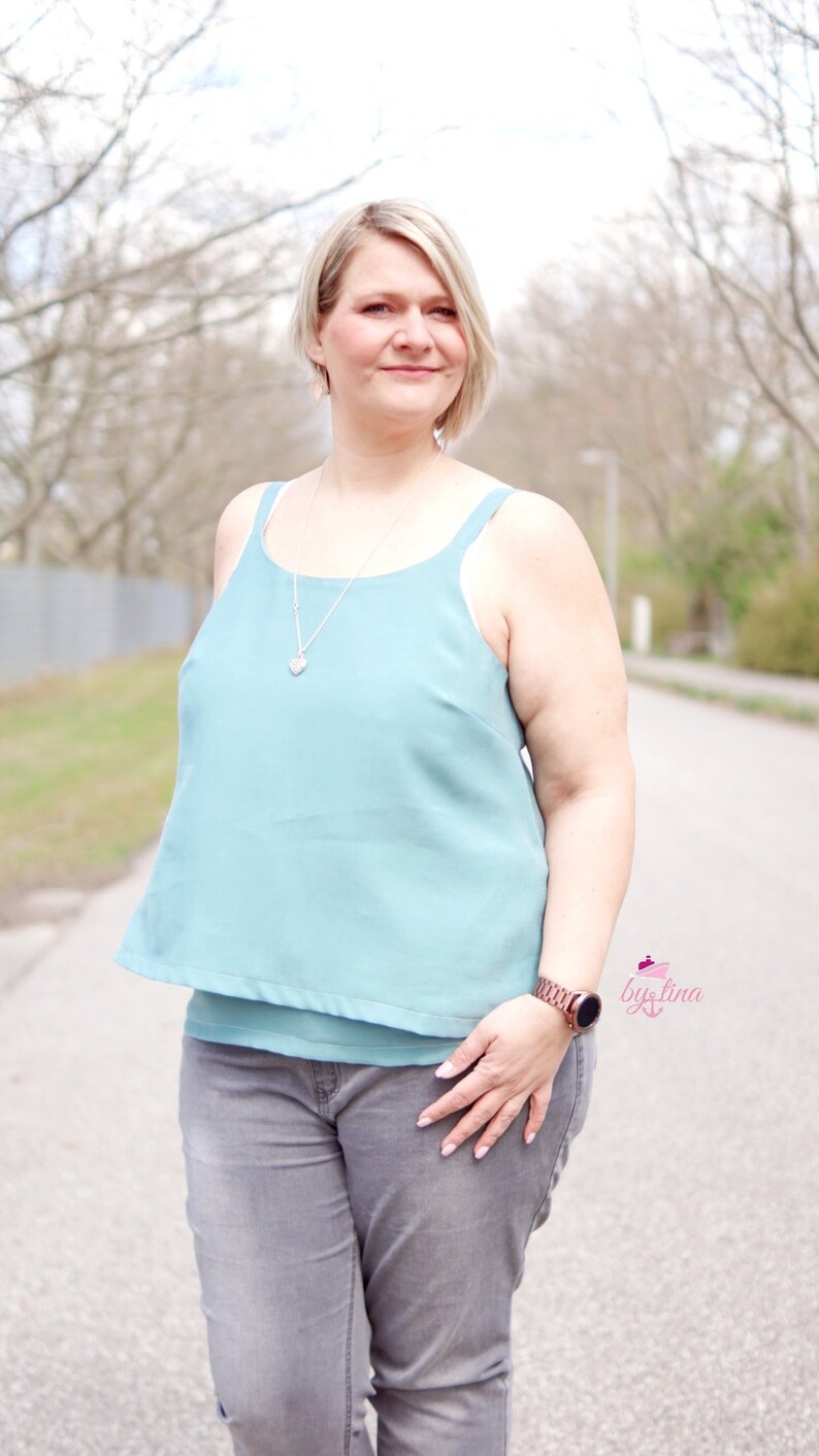 May include: A woman wearing a light blue sleeveless top with a silver necklace and a silver heart pendant. She is also wearing gray jeans and a brown watch.