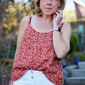 May include: A woman wearing a rust-colored tank top with a white floral pattern and white jeans. She is wearing a silver watch and glasses.