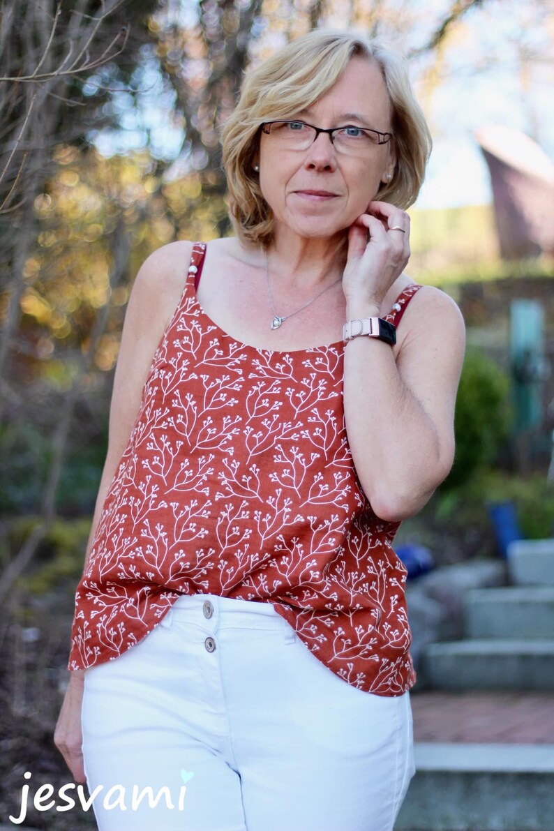 May include: A woman wearing a rust-colored tank top with a white floral pattern and white jeans. She is wearing a silver watch and glasses.
