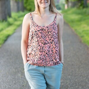 May include: A woman wearing a pink and black animal print tank top and light blue shorts. She is standing on a paved path with green trees in the background.