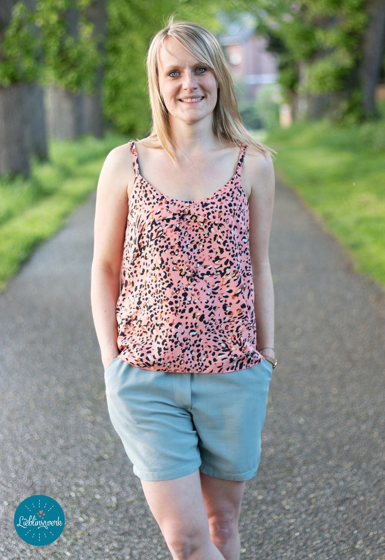 May include: A woman wearing a pink and black animal print tank top and light blue shorts. She is standing on a paved path with green trees in the background.