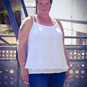 May include: A woman wearing a white, floral embroidered tank top with a blue denim skirt. She is standing in front of a metal fence.