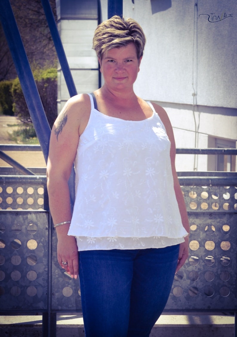 May include: A woman wearing a white, floral embroidered tank top with a blue denim skirt. She is standing in front of a metal fence.