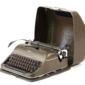 Könnte beinhalten: Eine olivgrüne Vintage-Schreibmaschine von Olympia mit passendem Tragekoffer. Die Schreibmaschine hat eine vollständige Tastatur mit dunklen Tasten und einen Chromwagen. Der offene Koffer zeigt die Schreibmaschine im Inneren.